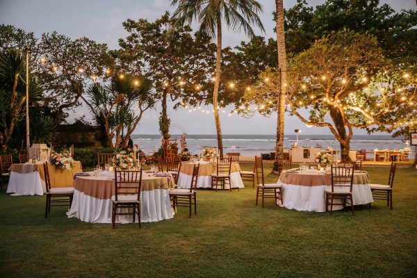 Wedding banquet or gala dinner decorated with garlands. Round tables styled with candles, flowers and accessories. Open air festive banquet on green lawn with sea view during dusk